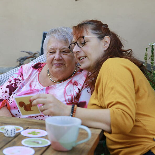Eine Betreuerin spielt geduldig mit einer demenzkranken Seniorin ein Kartenspiel im Garten.